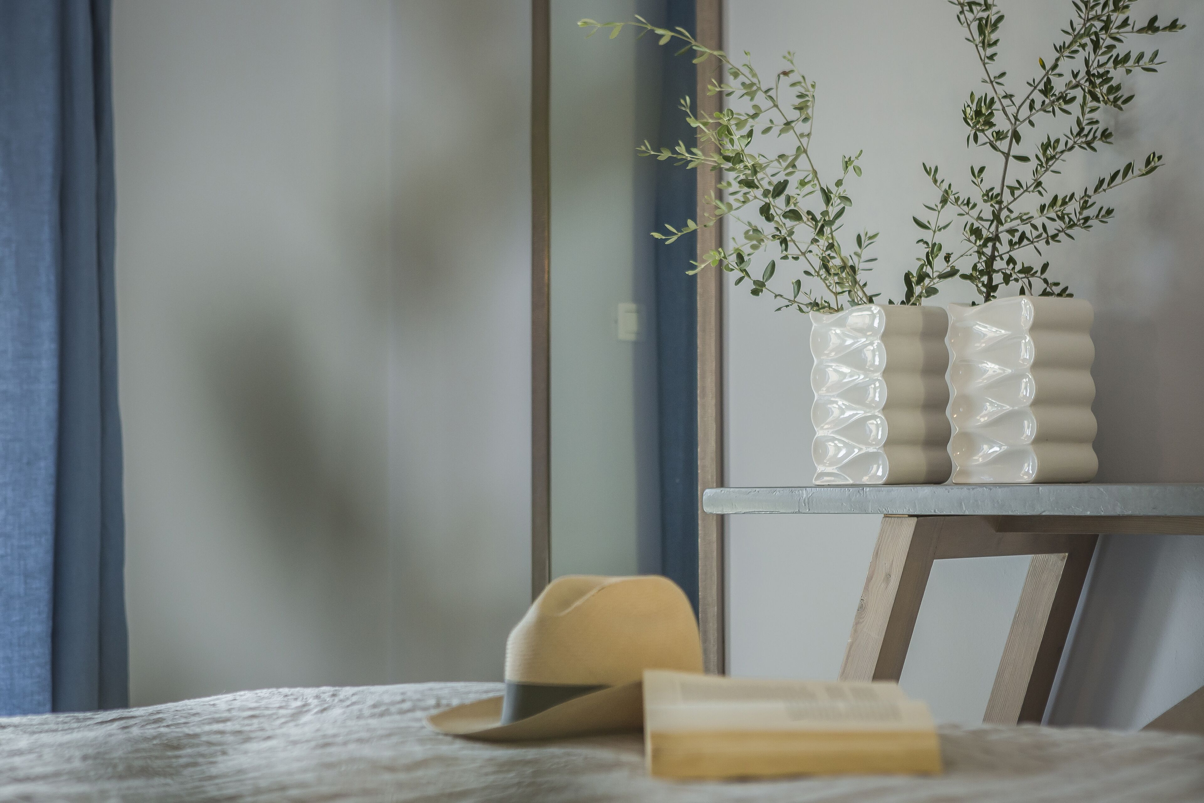 Una habitación con una cama, una mesita de noche de madera, un jarrón beige con una planta y un sombrero amarillo sobre la cama.