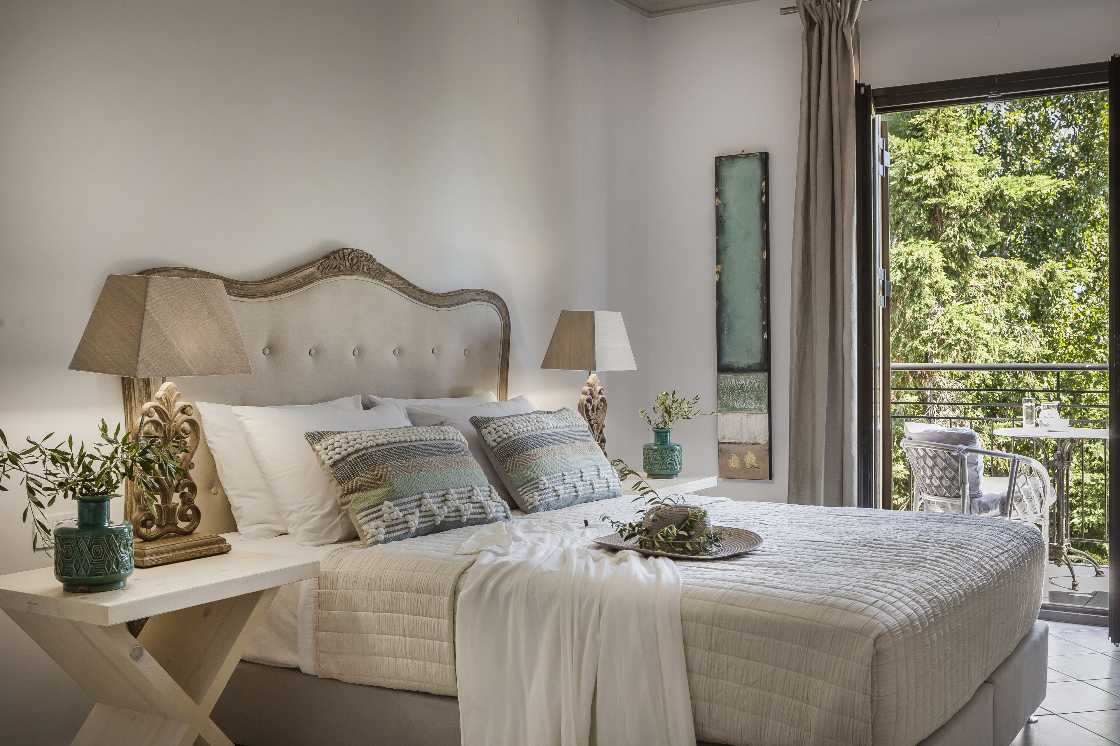 Une chambre avec un grand lit, deux tables de chevet avec des lampes, une vue sur la verdure par une porte-fenêtre donnant sur un balcon, et une tête de lit décorative.