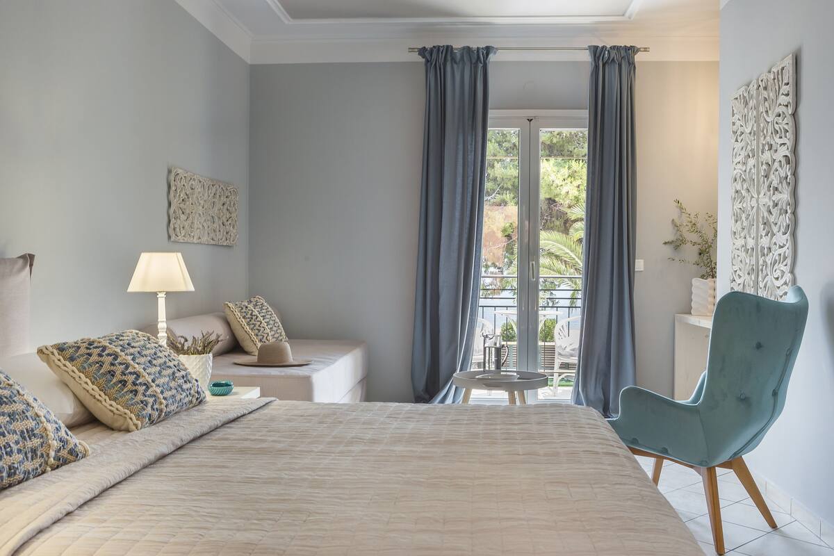 A bedroom with a large bed, a bedside table with a lamp, a chair, and a view of the outdoors through a window with curtains.