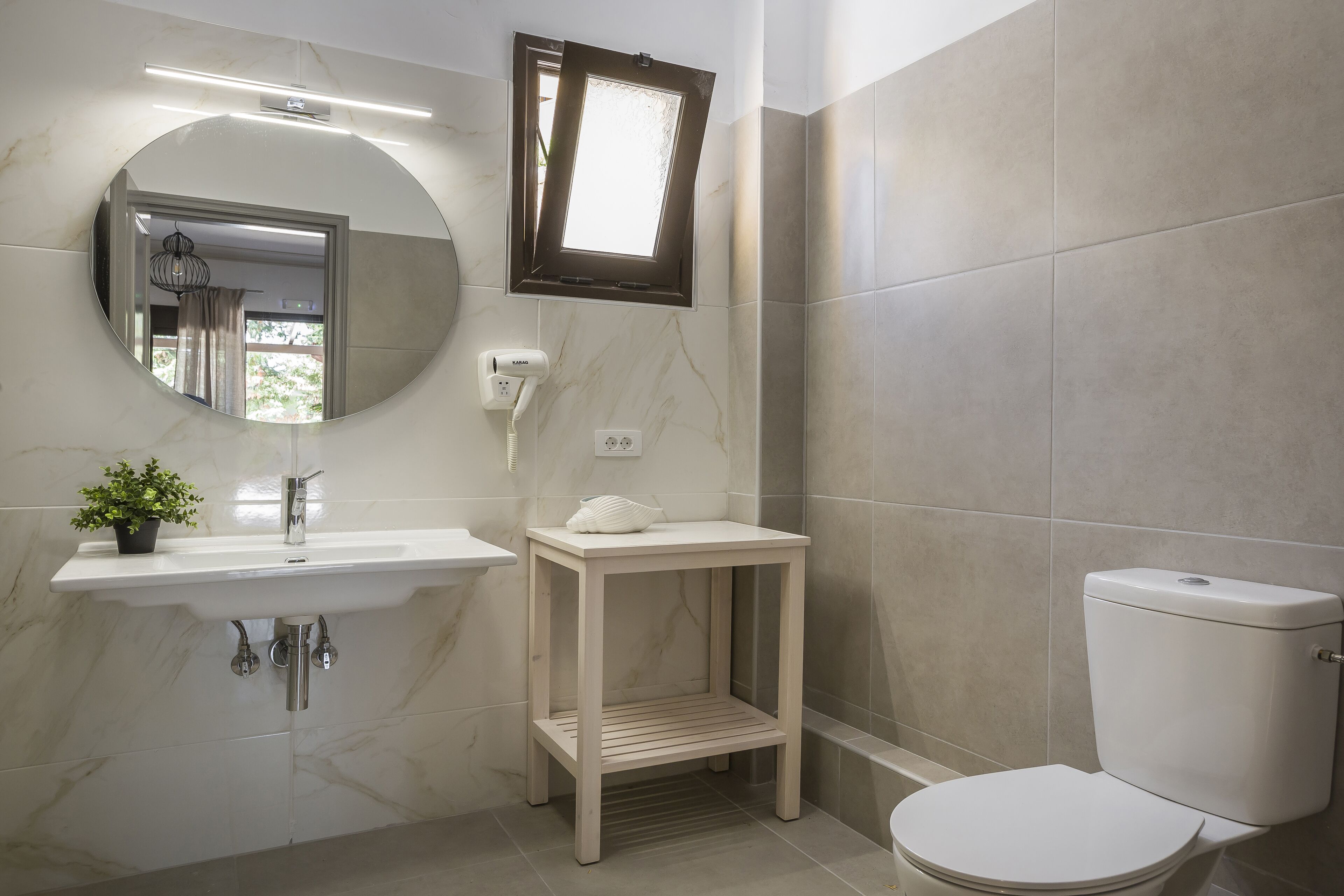 A bathroom with a round mirror, a sink, a wooden stool, and a toilet.