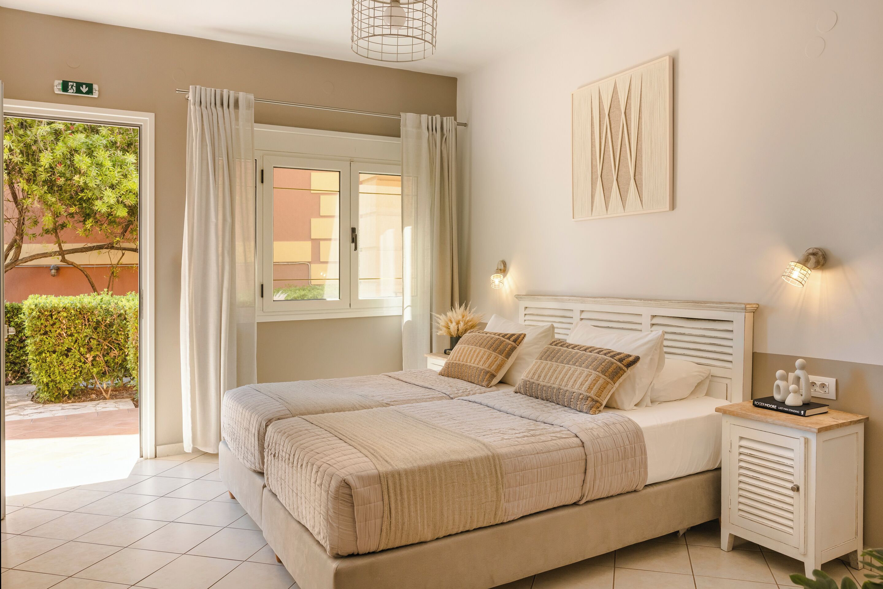 A bedroom with a large bed, a nightstand, a wall-mounted light, and a view of the outside.