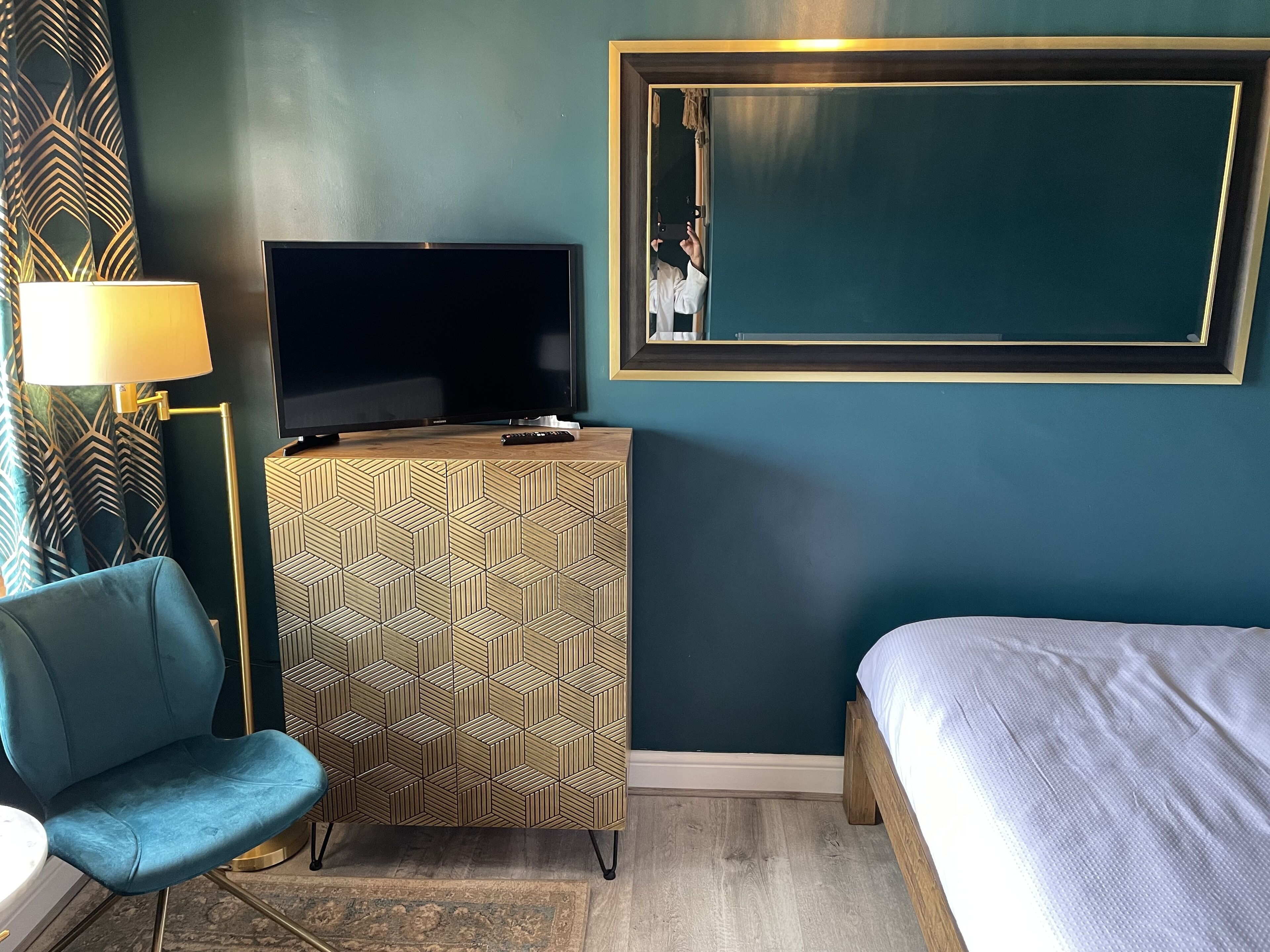 A hotel room with a TV on a wooden cabinet, a blue chair, a bed, and a large mirror on the wall.