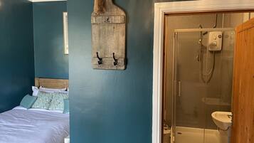 Ein Schlafzimmer mit einem Bett, einem an der Wand befestigten Holzregal und einem Badezimmer mit Duschkabine aus Glas und einem Waschbecken.