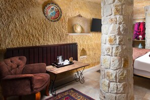 A cozy room with a stone wall, a wooden bench, a small table with a kettle, and a bed with a red blanket. - Romantic Cave Hotel (Ürgüp)