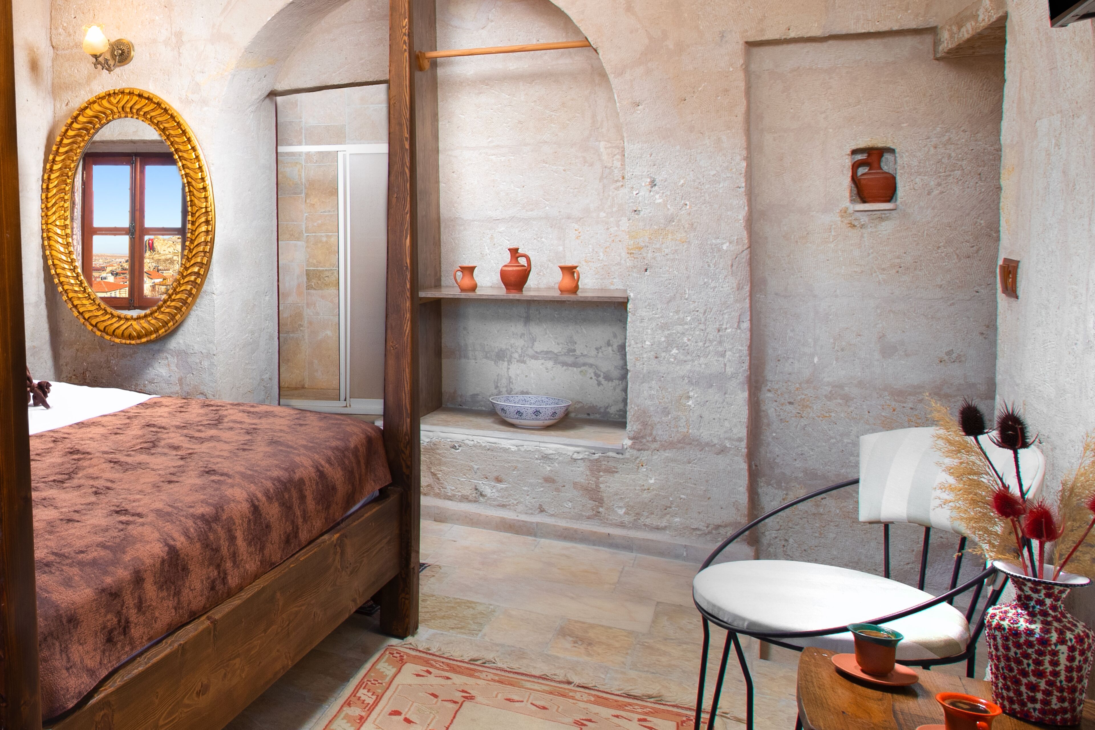 A rustic room with a bed, a mirror, a small table with a chair, and a view through an arched doorway.
