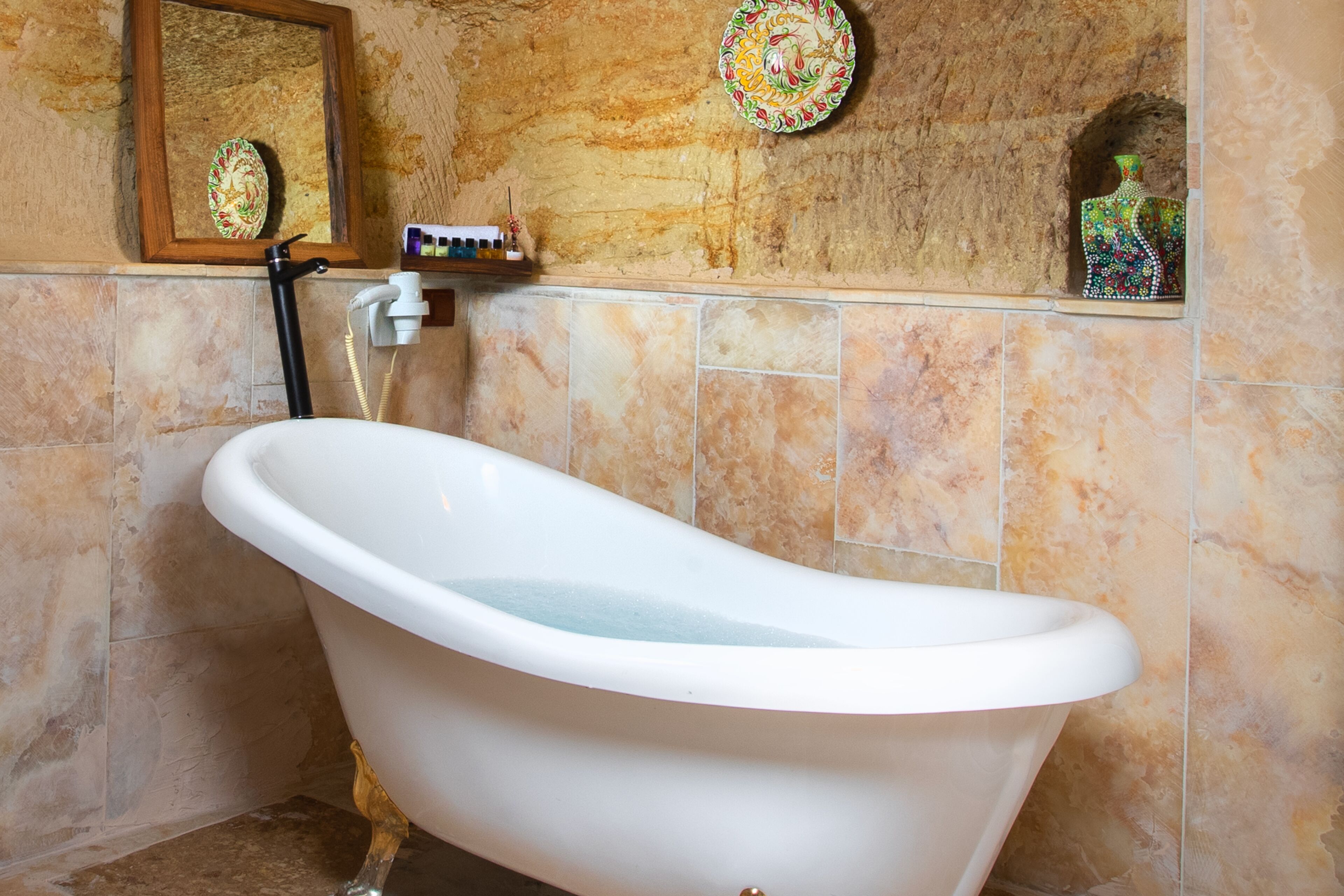 A white freestanding bathtub in a bathroom with tiled walls and a wooden shelf above it.