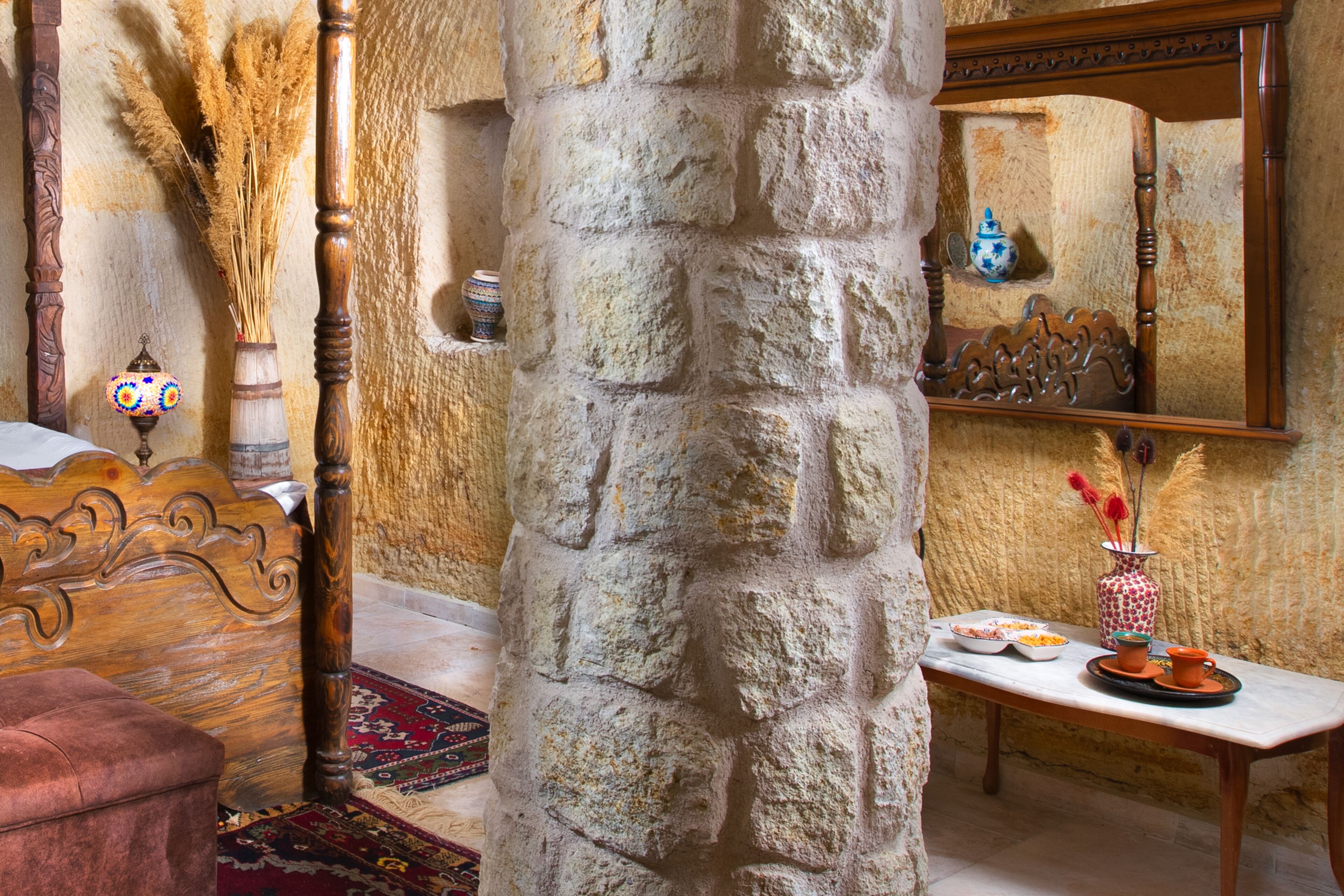 Una habitación con un pilar de piedra, una cama de madera, una mesita con un florero y un espejo en la pared.