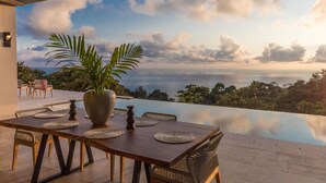 An infinity pool with a wooden table and chairs, a large potted plant, and a view of the ocean and sky. - Resol Luxury Oceanview Villa in Costa Rica (Bahía Ballena)