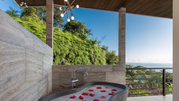 A stone bathtub with red flowers, a modern faucet, and a view of greenery and the sea. - Resol Luxury Oceanview Villa in Costa Rica (Bahía Ballena)