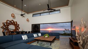 A modern living room with a large blue sofa, a wooden coffee table, and a view of the sunset over the ocean. - Luxe 4 Bdrm Villa w Epic Oceanview Infinity Pool (Bahía Ballena)
