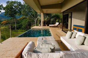 A modern outdoor pool area with lounge chairs and a sofa, surrounded by lush greenery and a mountain view. - Luxe 4 Bdrm Villa w Epic Oceanview Infinity Pool (Bahía Ballena)