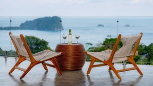 Deux chaises en bois, avec des assises et des dossiers en osier tressé, une table en osier, une bouteille de vin et deux verres à vin. La scène se déroule en plein air, avec une vue sur la mer et une île au loin.
