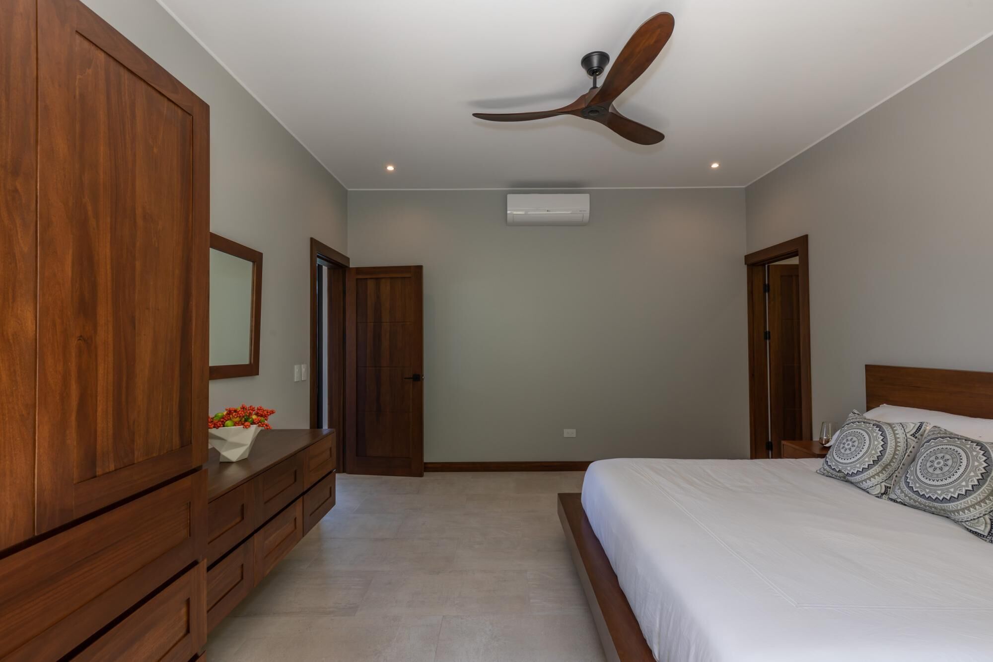 A bedroom with a wooden wardrobe, a bed with patterned pillows, a ceiling fan, and an air conditioning unit.