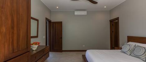 A bedroom with a wooden wardrobe, a bed with patterned pillows, a ceiling fan, and an air conditioning unit.