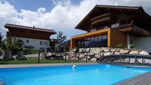 A swimming pool with a wooden deck, a stone wall, and a modern building with a balcony.