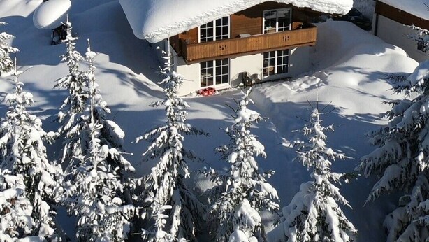 A snow-covered lodge with multiple wooden cabins, surrounded by snow-laden trees and mountains in the background.