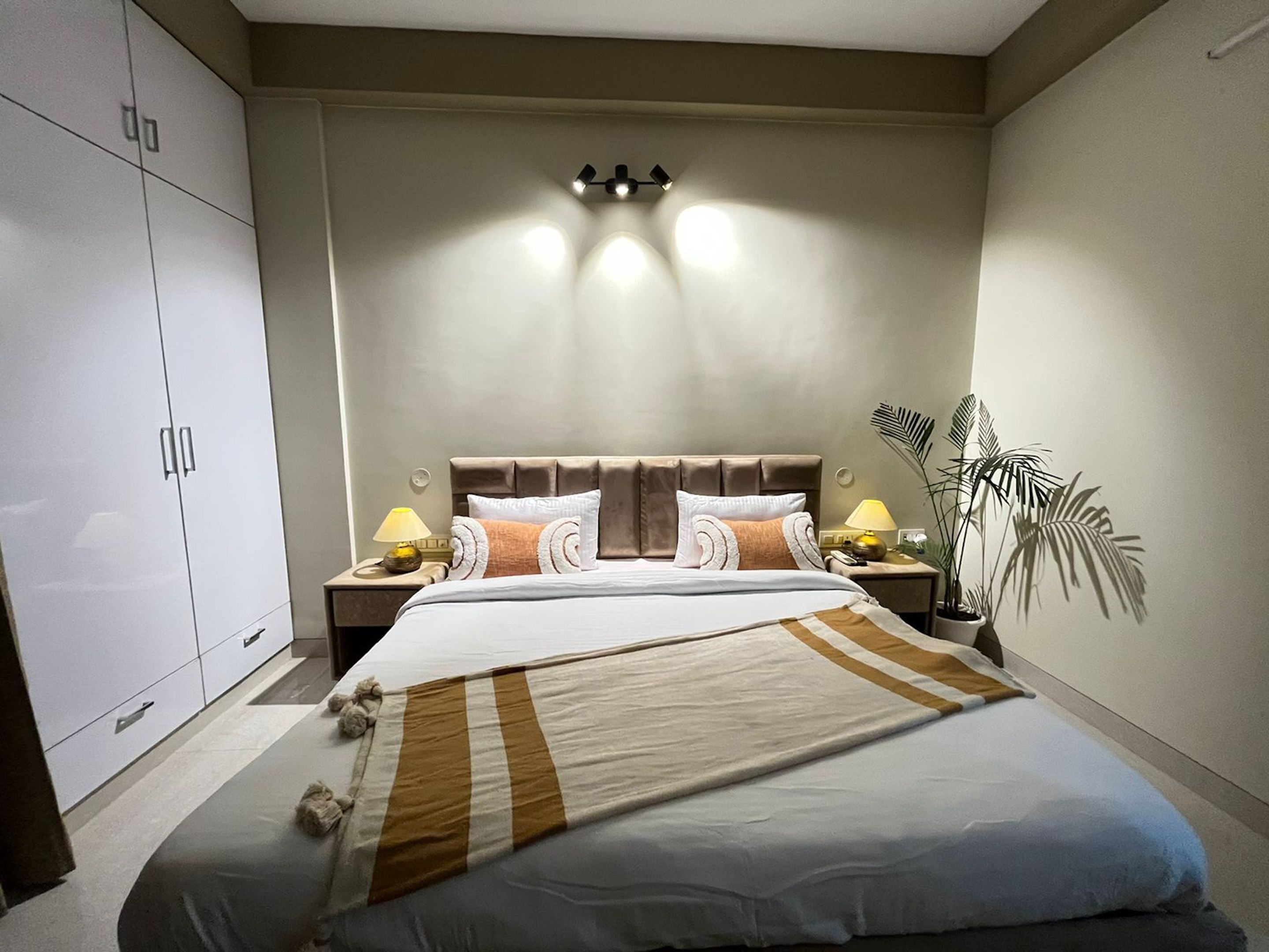 A bedroom with a large bed, two bedside tables with lamps, a wardrobe, and a potted plant.
