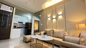 A modern living room with a sofa, coffee table, and a small kitchen in the background. - BedChambers Serviced Apartments Gurgaon (Gurugram)