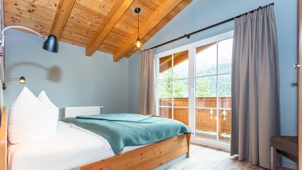 Ein Schlafzimmer mit einem Holzbett, einem Fenster mit Vorhängen und Blick auf Grün.