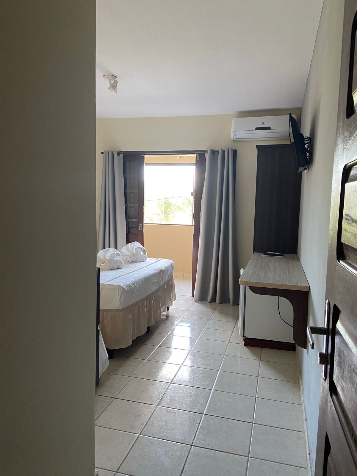 Ein kleines, sauberes Zimmer mit einem Bett, einem Schreibtisch und einem Fernseher. Es gibt ein Fenster mit Vorhängen und Blick ins Freie.