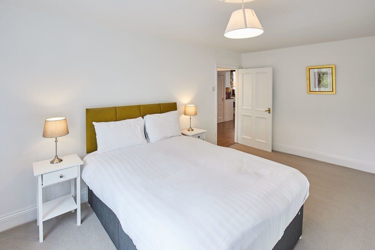 Un dormitorio ordenado con una cama grande, dos mesitas de noche con lámparas, una puerta que da a otra habitación y un cuadro enmarcado en la pared.
