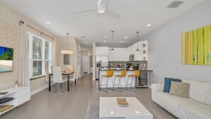 A modern living room with a white sofa, a coffee table, and a TV on the wall. There is a kitchen area with white cabinets and a dining area with yellow chairs. - 1511retreat 3 Bedroom Townhouse by Redawning (Clermont)