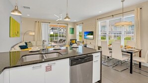 A modern kitchen with a dishwasher, dining area with white chairs, and a large window with blinds. - 1511retreat 3 Bedroom Townhouse by Redawning (Clermont)