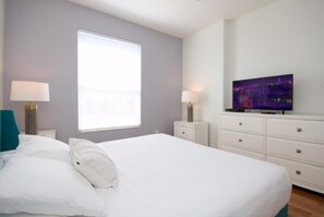 A bedroom with a bed, two bedside tables with lamps, a TV on a dresser, and a window with blinds.