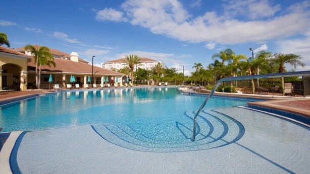 Uma piscina com espreguiçadeiras e guarda-sóis, rodeada por palmeiras e edifícios de hotel.