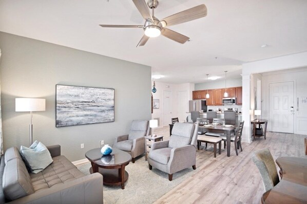 A modern living room with a sofa, armchairs, a coffee table, and a dining area with a table and chairs. There is a ceiling fan, a painting on the wall, and a kitchen in the background. - 3 Bed Penthouse Near Convention Center! 3 Bedroom Condo by RedAwning (Orlando)