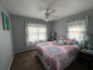 A bedroom with a bed, two windows with sheer curtains, a ceiling fan, and a nightstand with a lamp.