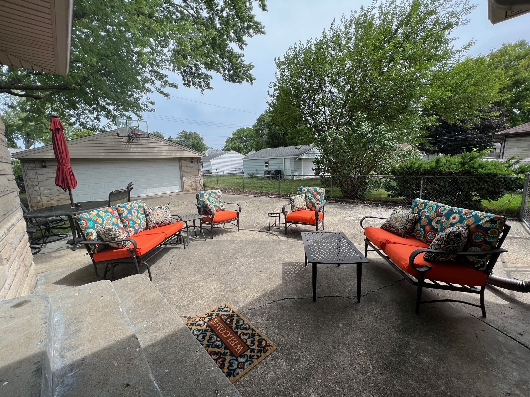 An outdoor patio with four chairs and a table, surrounded by a concrete floor and a welcome mat.