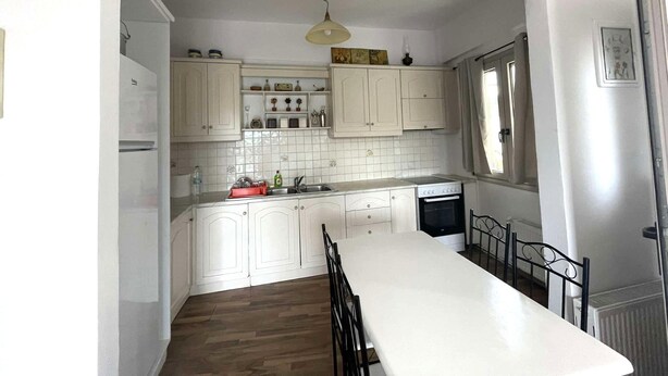A kitchen with white cabinets, a white countertop, and a dining table with chairs. There are framed pictures above the cabinets and a window on the right side.