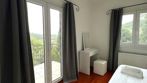 A hotel room with a wooden floor, a bed with white linens, a small white desk, a radiator, and a balcony with a view of greenery.