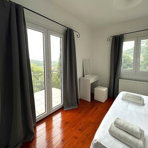 Et hotelværelse med trægulv, seng med hvide sengetøj, et lille hvidt skrivebord, en radiator og en balkon med udsigt over grøn bevoksning.