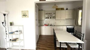 A kitchen with white cabinets, a dining table, and chairs. There is a shelf with books and a plant.
