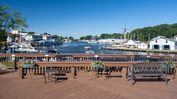 Une terrasse en bois, agrémentée de bancs et de jardinières, qui offre une vue sur une marina avec des bateaux et un ciel d’un bleu limpide.