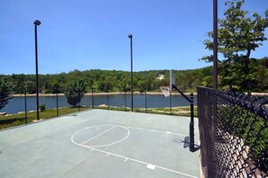 Un terrain de basket en plein air, équipé d’un panier, entouré d’une clôture en treillis métallique, avec un lac en arrière-plan.