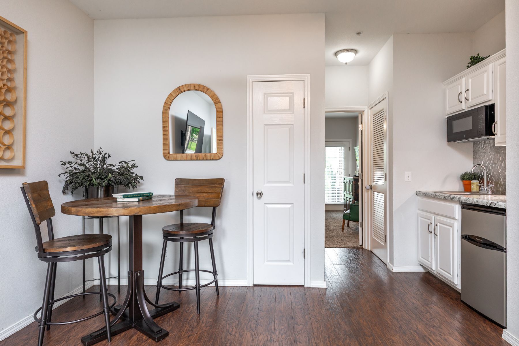 A dining area with a wooden table and two chairs, a white door, a kitchen with white cabinets and a stainless steel refrigerator, and a hallway leading to another room.
