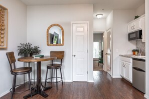 Un coin repas avec une table en bois et deux chaises, une porte blanche, une cuisine avec des meubles blancs et un réfrigérateur en acier inoxydable, et un couloir menant à une autre pièce.