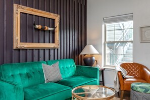 A room with a green sofa, a wooden frame with a horn, a lamp, and a window with a view of stairs.