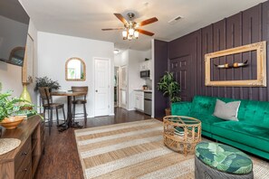 A living room with a green sofa, a dining area with a wooden table and chairs, a ceiling fan, and a kitchen in the background.