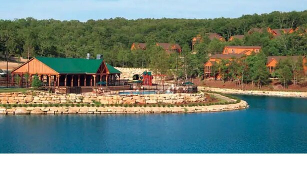 A lakeside resort with a large wooden building, a swimming pool, and a stone walkway.