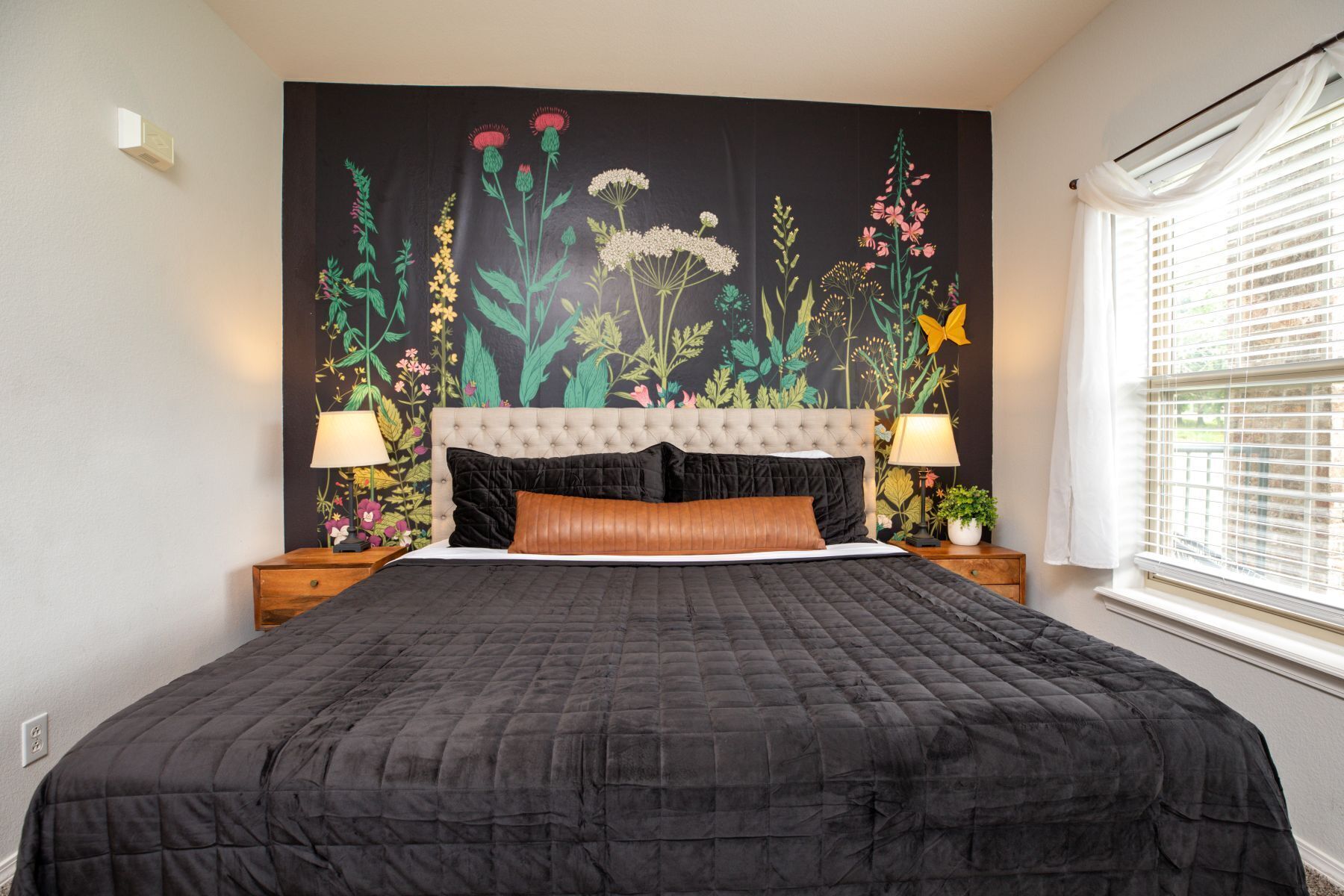 Une chambre à coucher avec un grand lit, une tête de lit capitonnée, deux lampes de chevet et une fresque murale à motifs floraux.