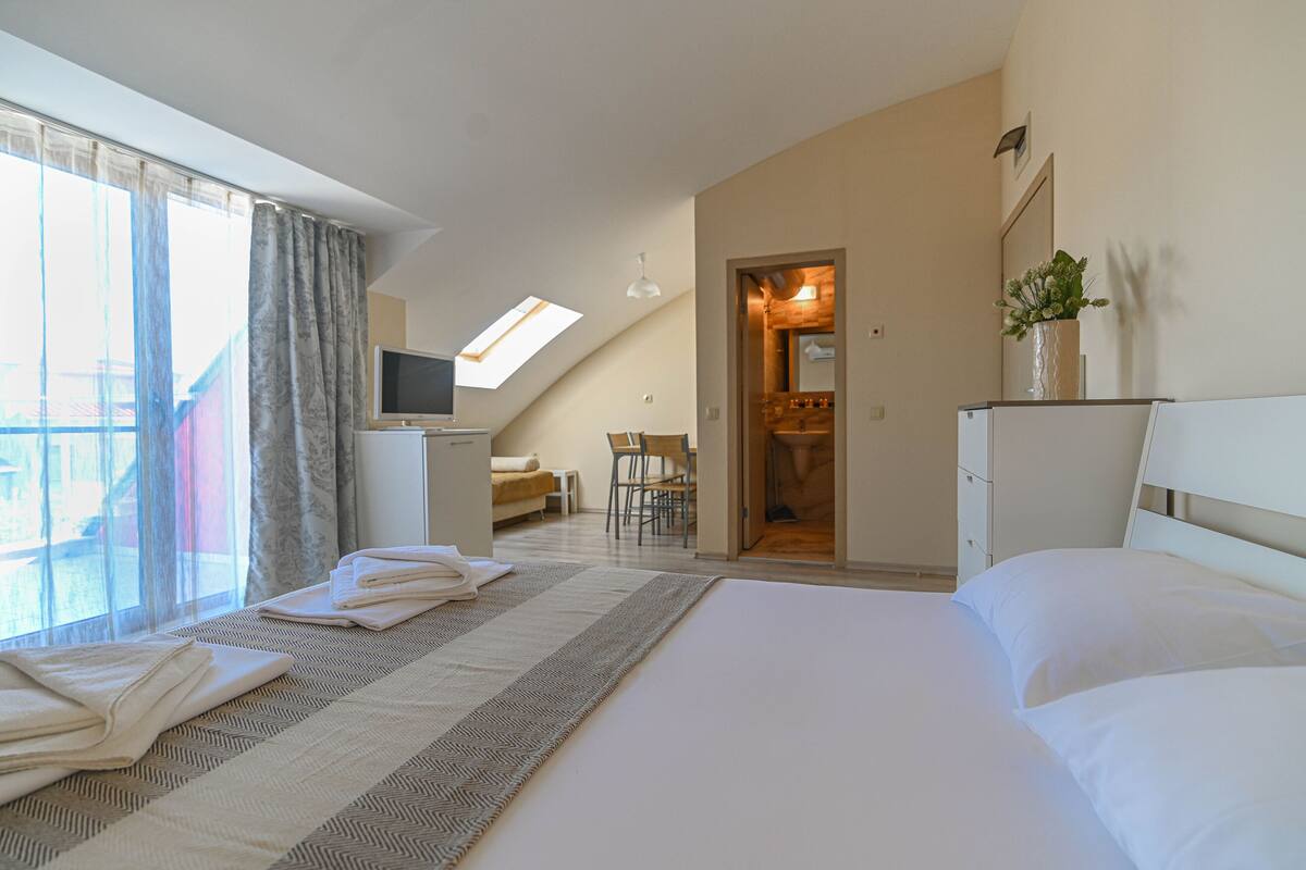 A hotel room with a bed, a desk, a chair, a TV, and a bathroom visible through an open door.