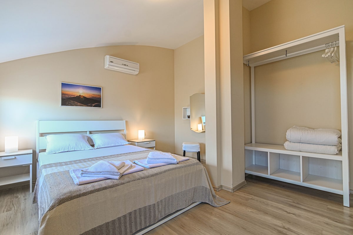 A neatly made bed with a patterned bedspread and pillows, a bedside table with a lamp, a wall-mounted air conditioner, a framed picture of a sunset, and a walk-in closet with folded towels.