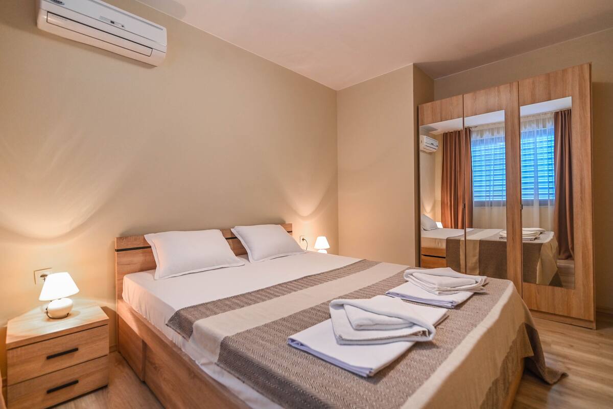 A hotel room with a bed, bedside tables, a lamp, an air conditioning unit, a mirror, and a wardrobe.