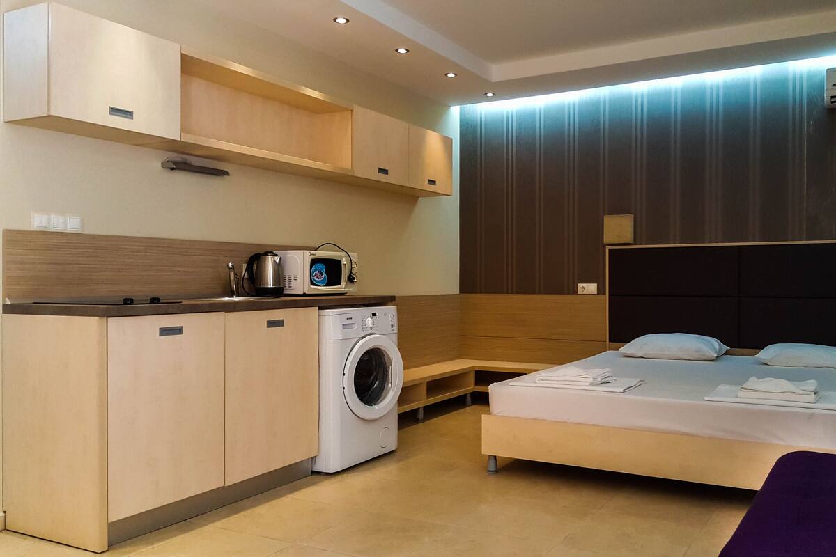 A modern hotel room with a washing machine, a microwave, a bed with pillows, and a purple chair.