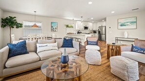 Un salon moderne avec un canapé, un pouf et une table basse, un coin repas avec des chaises, et une cuisine en arrière-plan.