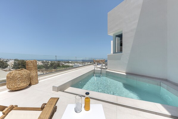 A rooftop pool with clear blue water, surrounded by white walls and a glass railing. There are wicker chairs and a table with a glass and a bottle.